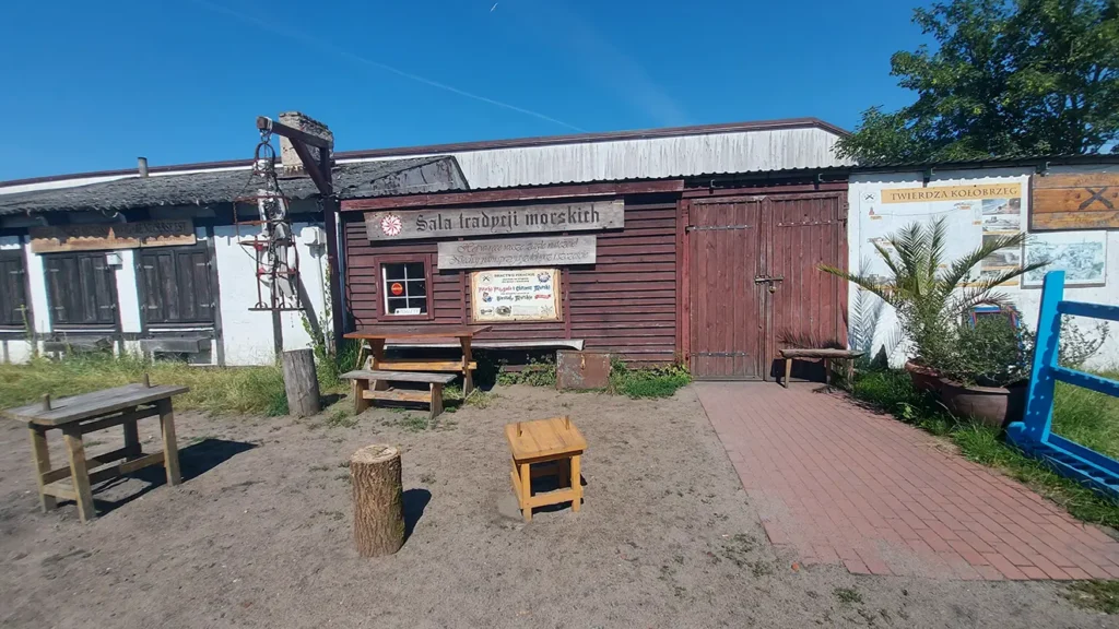 Kołobrzeska Sala Tradycji Morskich. Widać wiszącego kościotrupa, stoły, palmy, ławki. Na ścianie budynku jest duży napis z różą wiatrów "Sala Tradycji Morskich".